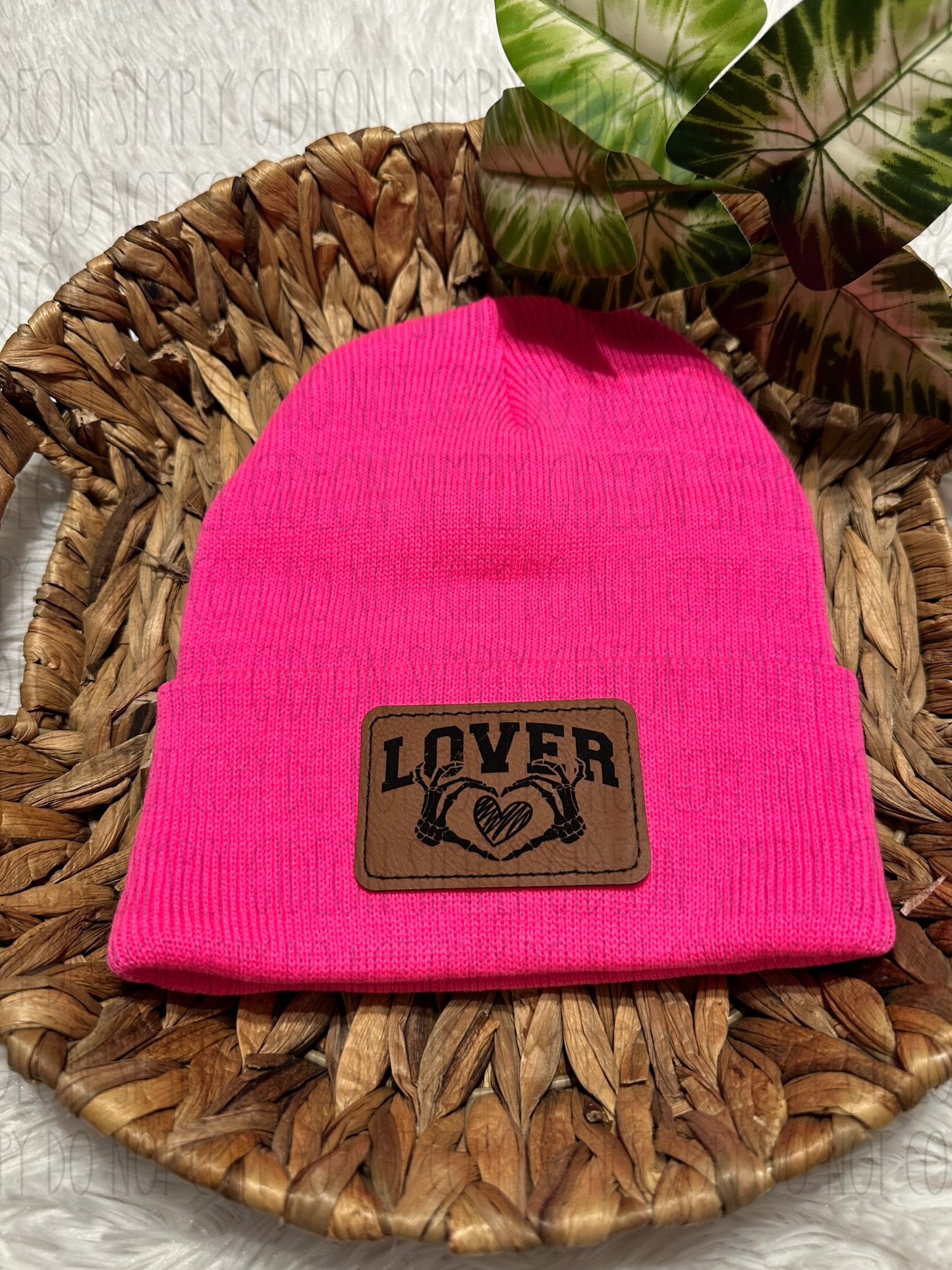 A neon pink acrylic beanie with a brown laser engraved patch that has an image with skeleton hands making a heart with a heart inside and reads lover