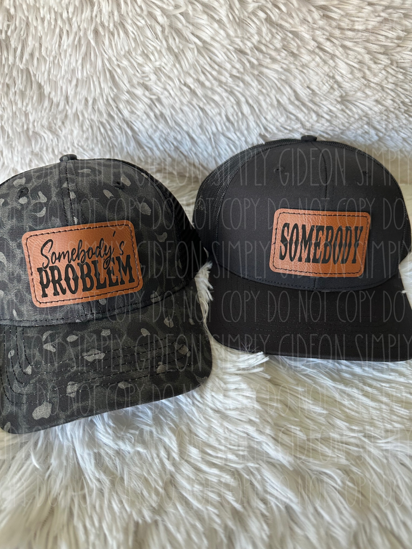 two hats one black and grey leopard print hat with a laser engraved patch that reads somebody's problem and a black hat with a brown laser engraved patch that reads somebody